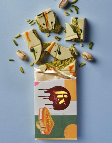 Colorful pistachio chocolate bar wrapper with pieces of white chocolate, pistachio pieces, and whole pistachios scattered on blue background